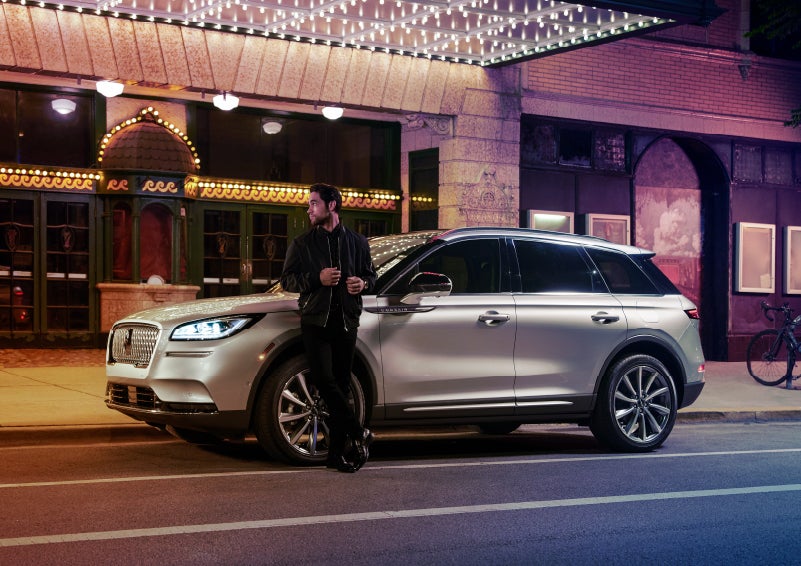 A 2022 Lincoln Corsair SUV is parked outside a theater as the driver relaxes against the frame and lights illuminate the floating roofline and body | Asheville Lincoln in Asheville NC