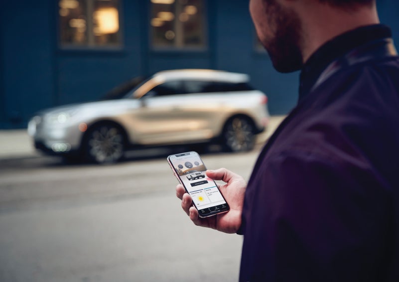 A man is holding his phone looking at the Lincoln Way™ App and we see his 2022 Lincoln Corsair SUV in the background against a blue brick wall | Asheville Lincoln in Asheville NC