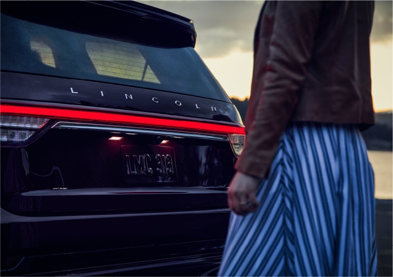 A person is shown near the rear of a 2023 Lincoln Aviator® SUV as the Lincoln Embrace illuminates the rear lights | Asheville Lincoln in Asheville NC