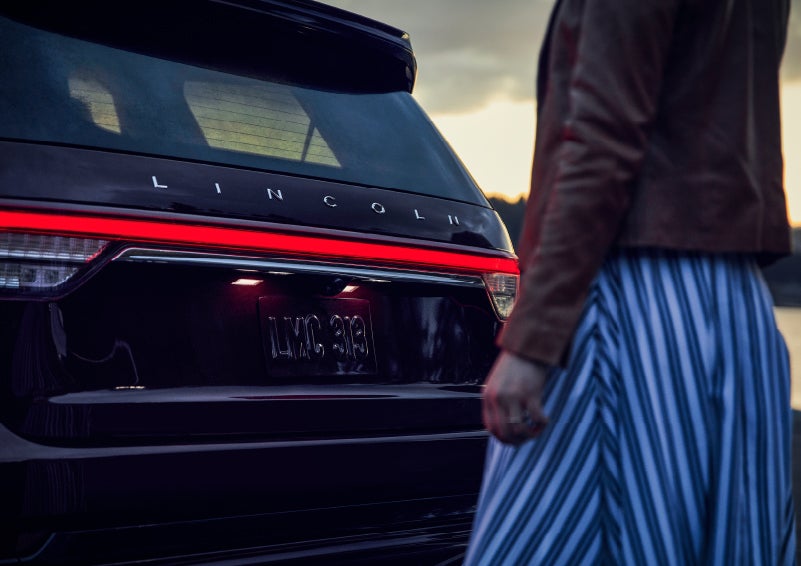 A person is shown near the rear of a 2024 Lincoln Aviator® SUV as the Lincoln Embrace illuminates the rear lights | Asheville Lincoln in Asheville NC