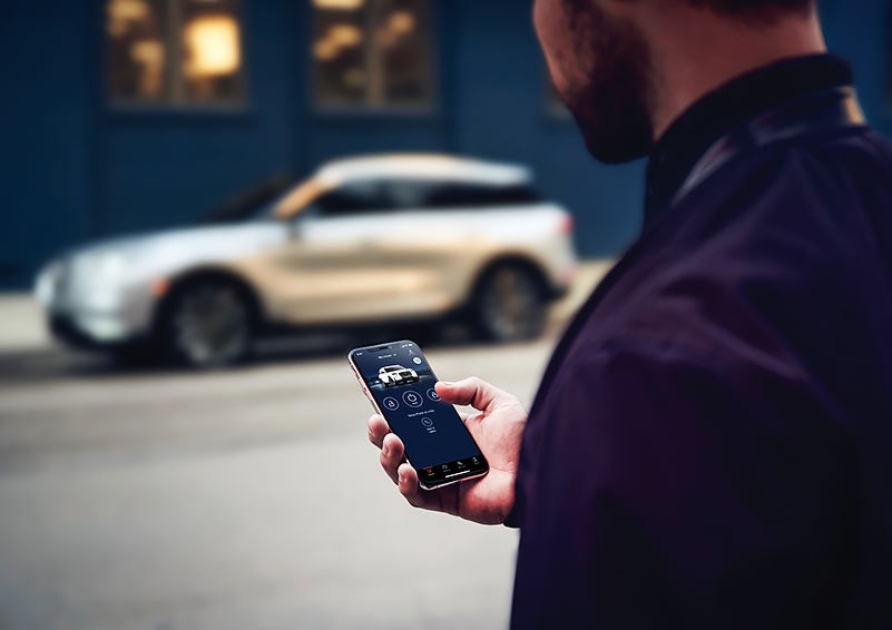 A person is shown interacting with a smartphone to connect to a Lincoln vehicle across the street.